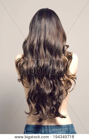 Rear view of a topless woman with long brown wavy hair against gray background