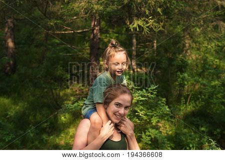 Mother and daughter in the forest. The little girl is sitting on her mothers' shoulders. The harsh sunlight is shining right in her face and she is screwing up her eyes