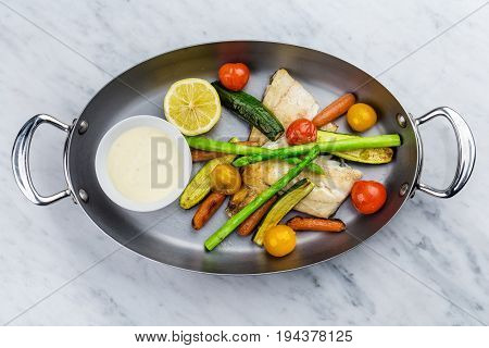 Grilled fish and vegetables with sauce served in metalic bowl on marble serface