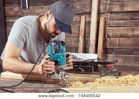A young man in a gray cap and a gray T-shirt carpenter builder holds a milling machine in the hand and equips the edges of the wooden board in the workshop wooden sawdust flies to the sides