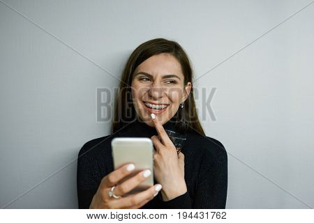 Headshot of attractive cheerful young female with long dark hair and braces texting messages using online messenger on her mobile phone having flirty look touching lips and smiling broadly