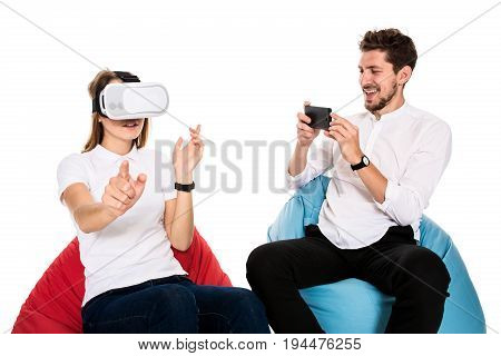 Smiling friends experiencing virtual reality glasses seated on beanbags isolated on white background. Experiencing virtual gaming adventure.