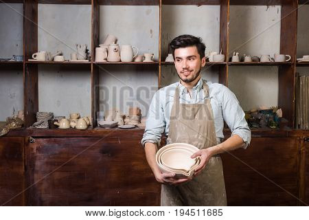 potter, tools, ceramics art concept - handsome young brunette male with a beard dressed in an apron stand with finished utensils, smiling ceramist at workplace with baked cups and jugs at background