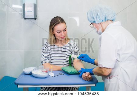Preparation for blood test with pretty young blond woman by female doctor in white coat medical uniform on the table in white bright room. Nurse pierces the patient's arm vein with needle blank tube
