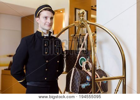 Smiling man in bellboy uniform standing with luggage cart and looking away.