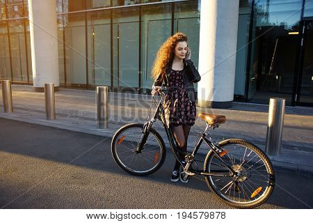 Cute teenage girl wearing stylish clothing standing on desert street with her vintage bike and talking on mobile phone. Pretty woman making stop answer phone call while riding bicycle on city streets