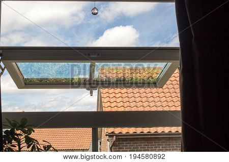 Urban living. View from open UPVC window reflecting garden on modern urban housing estate. Blue cloudy sky and roof tops visible through house window on hot day.
