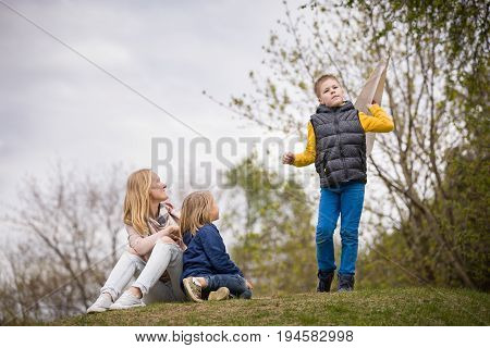 Portrait of happy family having fun outdoors. Mother with two boys. Kid starting a paper airplane and his brother and mom watching this while sitting on the grass in the park. Lifestyle.