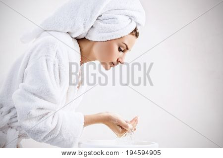 Young woman washing face with clean water in white bathrobe and towel. Morning hygiene concept.