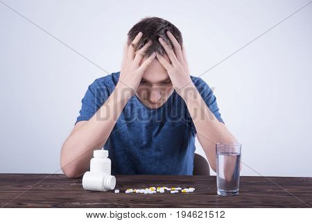 A young guy in a blue T-shirt is sitting at a table and holding his head in his hands. Pills and a glass of water are on a wooden brown table. The guy drinks pills. Healthy generation. Healthy youth.