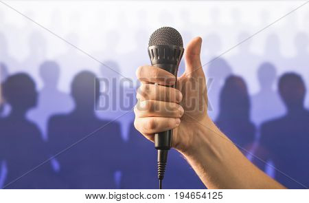 Hand holding microphone and showing thumbs up in front of a crowd of silhouette people. Public speaking and giving speech concept. Singing well live to mic, good karaoke or successful talent show.