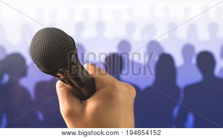 Public speaking and giving speech concept. Close up of hand holding microphone in front of a crowd of silhouette people. Singing to mic in karaoke or talent show concept.