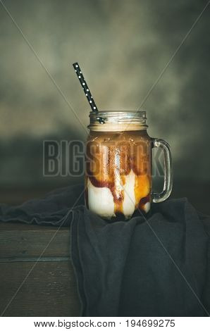 Iced caramel macciato coffee with milk in glass jar with straw on dark rustic wooden table, selective focus, copy space. Cold sweet refreshing summer drink concept