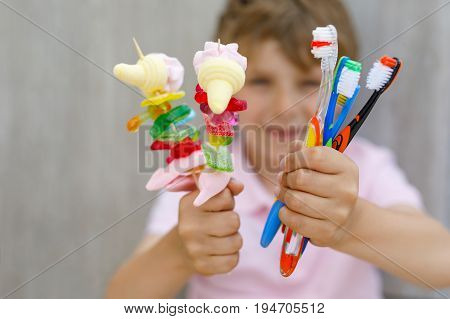 Happy little kid boy holding marshmallow skewer in one hand and toothbrushes in another. Child with different unhelthy colorful sweets. Healthy food and teeth concept.