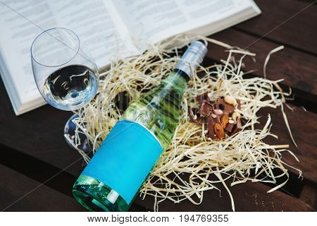 Glasswine with white wine, bottle and pieces of chocolate lies on sawdust near big open book, romantic background with expensive alcoholic drink