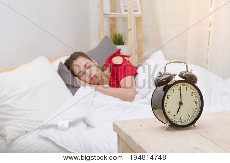 asian young woman in red pajamas sleeping on bed and black vintage retro alarm clock times at 7 o'clock on wooden desk in bedroom at the morning selective focus holiday lifestyle concept
