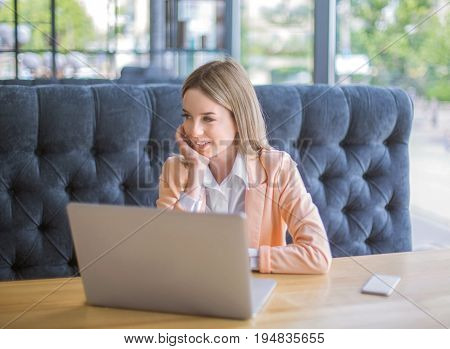 Happy female sits on the sofa and looks away. Portrait of beautiful, successful and smiling business woman in white blouse. The laptop and phone are on a light brown table.
