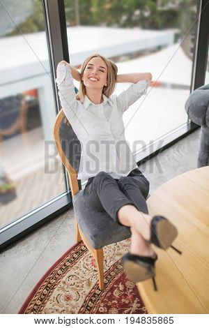 Gorgeous young happy female blond sits on the chair and holds her hands behind her head. A successful business woman is smiling. Full length image of young and slim business woman in the office.