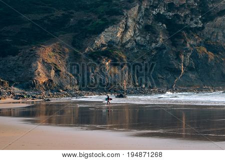 Very wide and open beach with tide line and mountain rock surfer walks towards waves with his surfing board ready to take on adventures