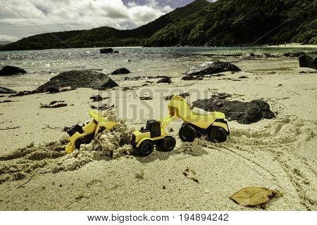 Toy construction trucks moving sand around on the beach of a tropical island, including a toy bulldozer, front loader, and dump truck