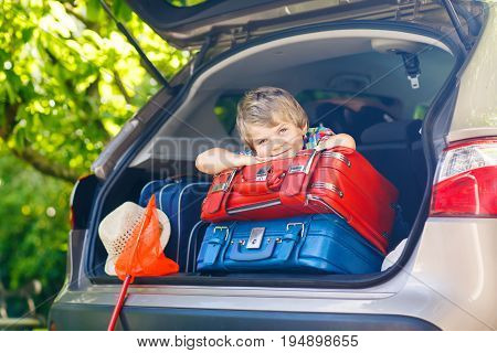 Adorable little kid boy sitting in car trunk just before leaving for summer vacation with his parents. Happy child with suitcases and toys going on journey. happy family traveling. Summer travel.