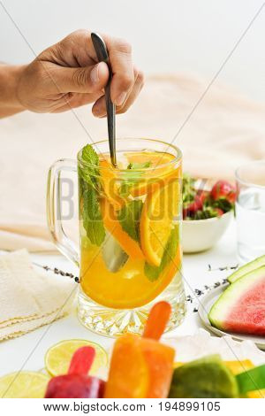 young man stirring a jar with a refreshing drink, with orange slices and mint, placed on a table next to a bowl with strawberries, a plate with watermelon and a plate with different homemade ice pops