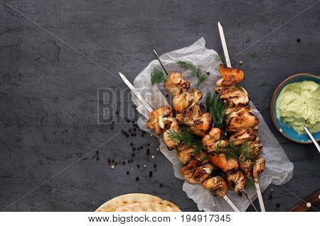 Barbecued chicken breast skewers with flat bread and avocado sauce on dark background with copy space top view