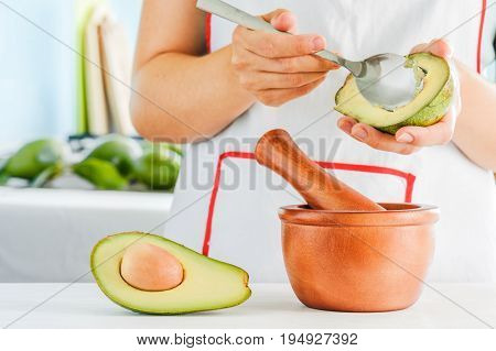 Woman making traditional Mexican sauce Guacamole from fresh ripe avocados. Wooden mortar with pestle (molcajete) and half of avocado on kitchen table. Healthy homemade vegetarian eco food.