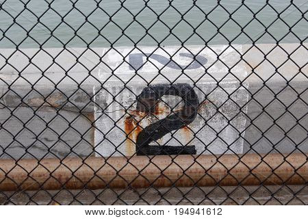 Looking through a black chain link fence at the number 2 painted in black against a white background on a conrete wall