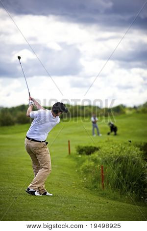 Golfspiller pitching golfbolden over divot med anden spiller i baggrunden ser ham.