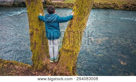 Young Boy Standing On The Tree And Enjoying The Picturesque View Of The River.