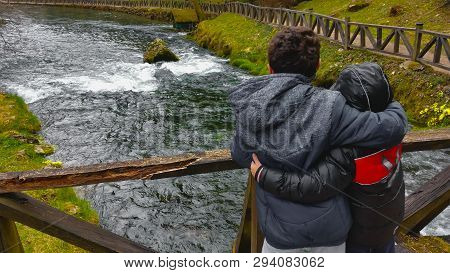 A Young Man Hugged With His Brother Stands On A Wooden Bridge And Enjoys A Picturesque View Of The R