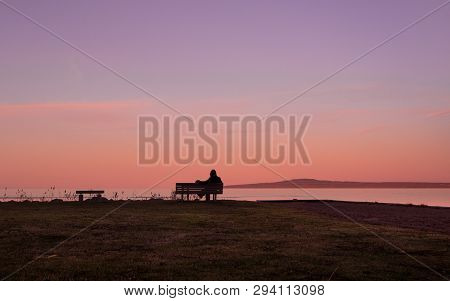 Man Sitting On Parkbench Looking Out Over The Water In Nature Twilight Sky