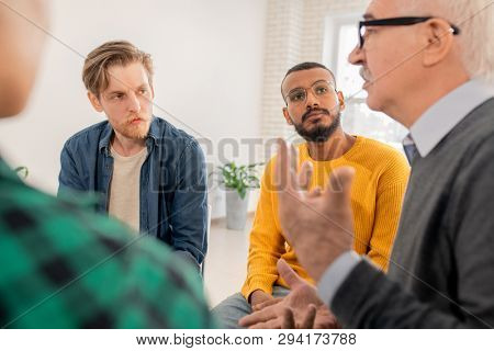 Two young intercultural men looking at senior groupmate talking about his problem at psychotherapy session