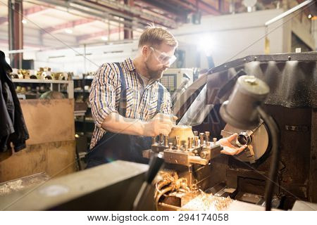 Young mechanic in workwear snd protective eyeglasses standing by huge industrial machine and preparing it for work