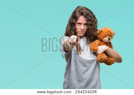Young brunette girl holding teddy bear over isolated background pointing with finger to the camera and to you, hand sign, positive and confident gesture from the front