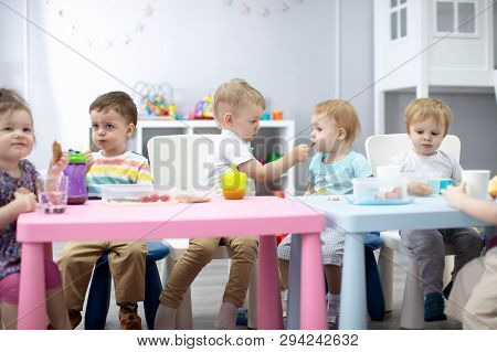 Kids Group Have Lunch In Kindergarten. Cute Child Boy Sharing His Food To Little Girl