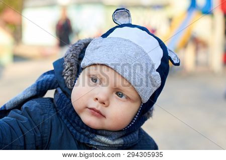 Portrait Of A Cute Little Blue-eyed Boy With An Interested Look In A Hat, Scarf And Jacket In Early