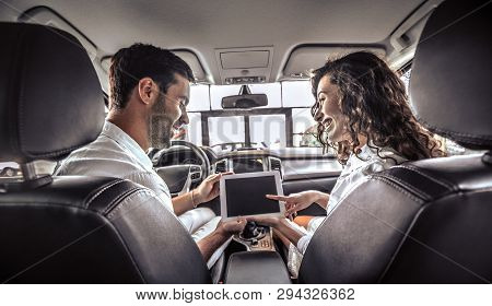 Couple Is Looking On Tablet In A New Car. The Are Looking Options Of Brandnew Car. Young Family Want