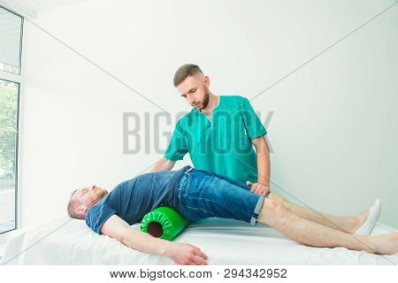 Man At The Physiotherapy Doing Physical Exercises With His Therapist, They Using A Massage Roll. A C