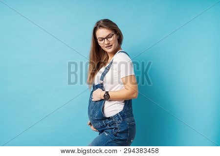 Pregnant Woman Caressing Her Belly Over Blue Background. A Cute Young Woman Wearing Glasses, And Den