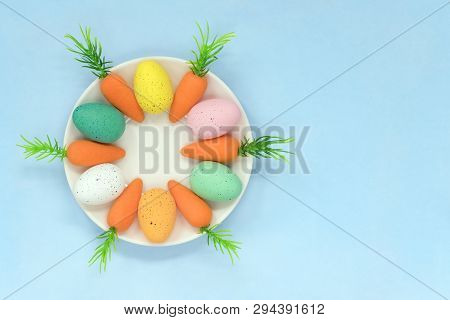 Colourful Easter Eggs And Carrots Placed Around A Saucer, On A Light Blue Background With Copy Space