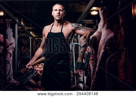 Strong Butcher With A Tattoo On His Hand Wearing Apron Posing With Two Axes In A Refrigerated Wareho