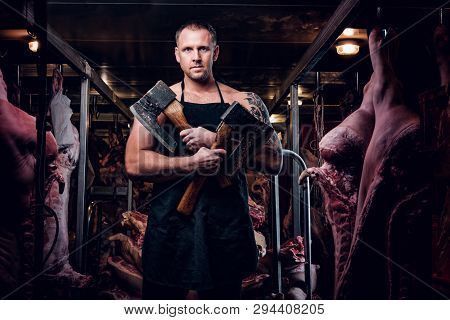 Shirtless Butcher With A Tattoo On His Hand Wearing Apron Posing With Two Axes In A Refrigerated War