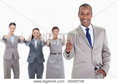Happy businessman with a hand in his pocket and approving with co-workers in the background with thumbs up