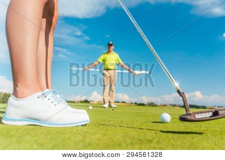 Golf ball on the putting green in selective focus behind the low section of a female player during class, with an experienced teacher on professional ground