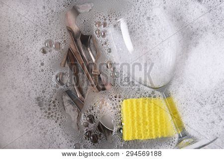 Overhead shot of a dishpan filled with soapy water with cutlery, a wine glass and sponge. 