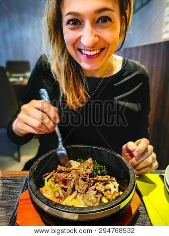 Korean Bibimbap Served In Hot Stone Dolsot Pot Bowl Eaten By Caucasian Smiling Woman Dining In Korea