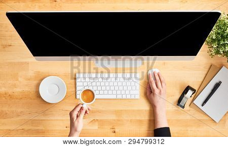 Female Hands Working On Modern Desktop Computer And Holding Cup Of Coffee. Top View Of Wooden Office