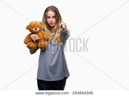 Young beautiful blonde woman holding teddy bear plush over isolated background pointing with finger to the camera and to you, hand sign, positive and confident gesture from the front